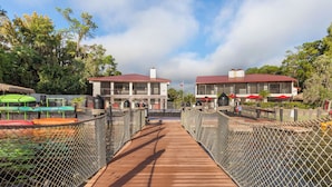 A wooden bridge leading to a building with a red roof, surrounded by greenery and a clear sky.