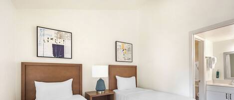 A hotel room with two beds, a wooden headboard, a nightstand with a lamp, and a bathroom visible through an open door.