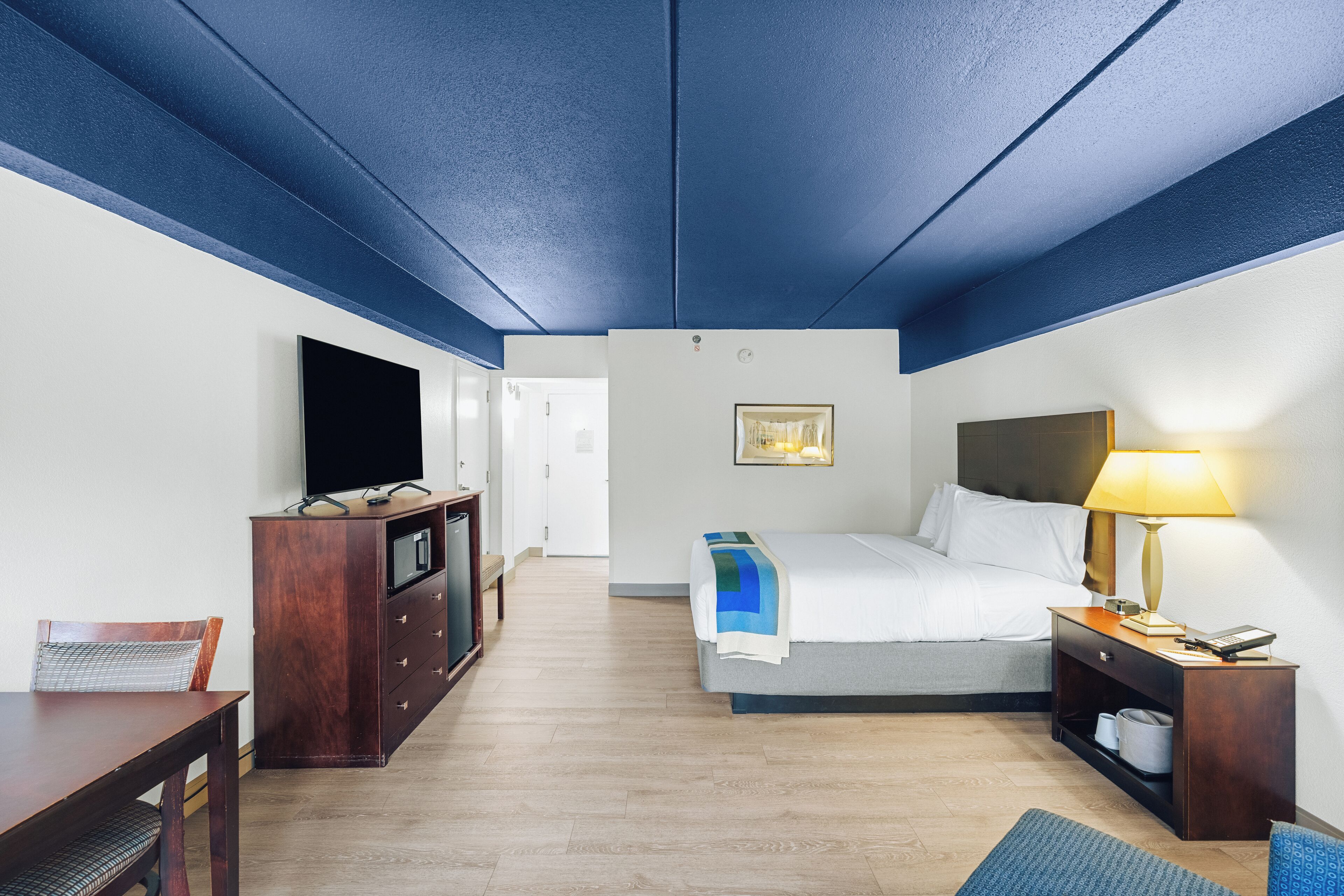 A hotel room with a bed, bedside table, lamp, TV, and a desk with a chair.