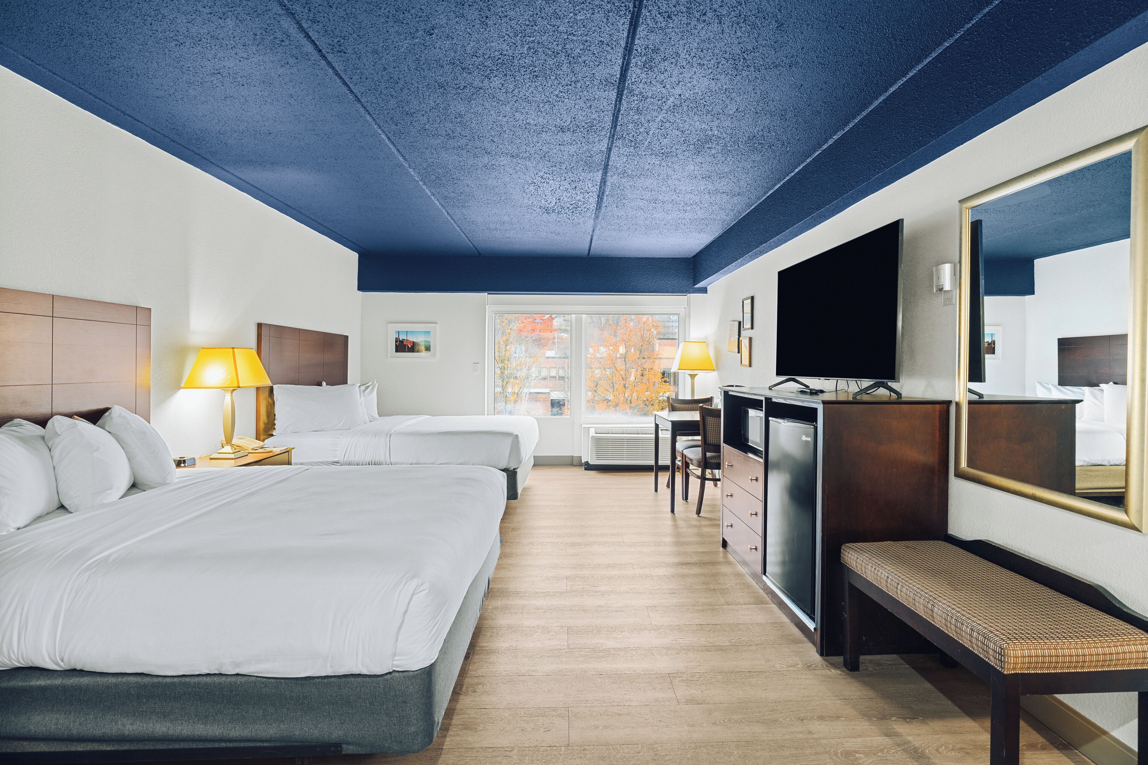 A hotel room with two beds, a desk, a TV, and a bench.