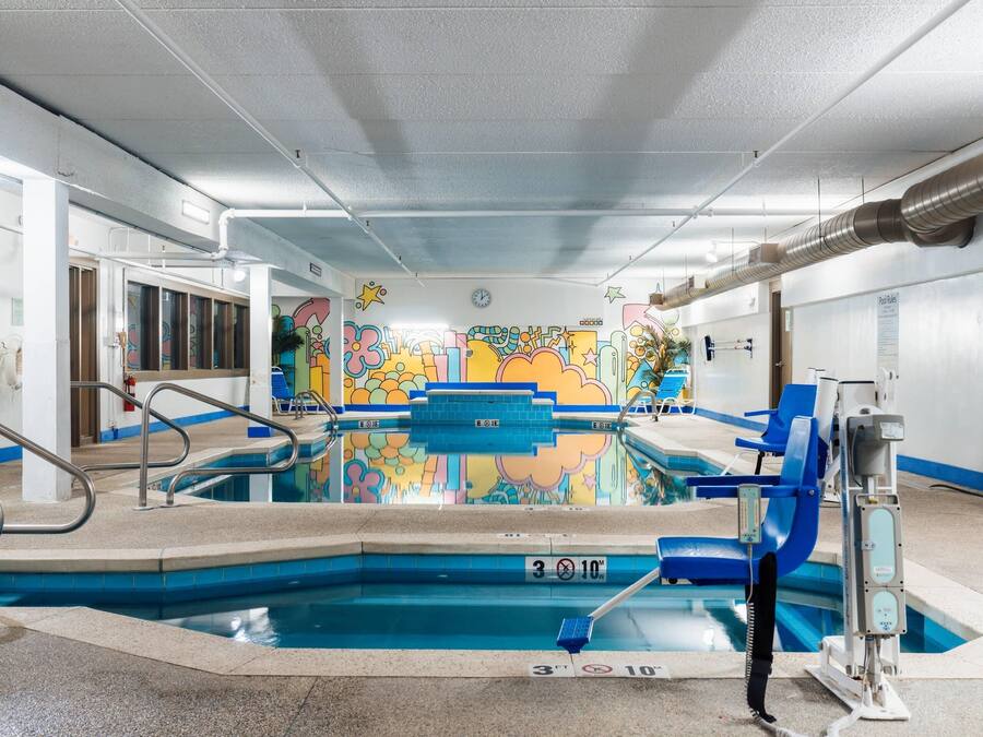 Piscina interior con un mural colorido en la pared, un puesto de socorrista y normas de la piscina visibles.