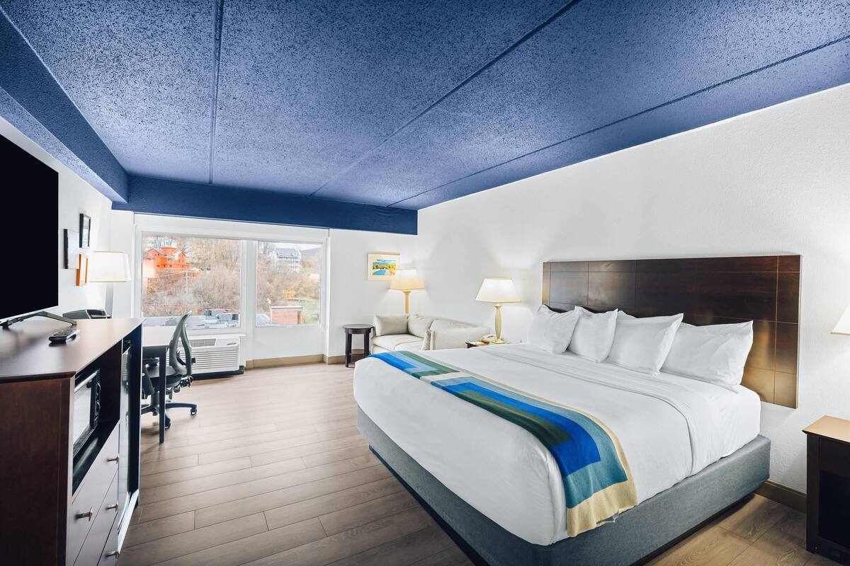 A hotel room with a large bed, a desk with a computer, a chair, and a window with a view of buildings outside.