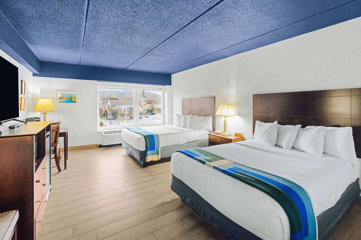 A hotel room with two beds, a desk, and a chair. There is a window with a view of buildings and a door leading to another room.