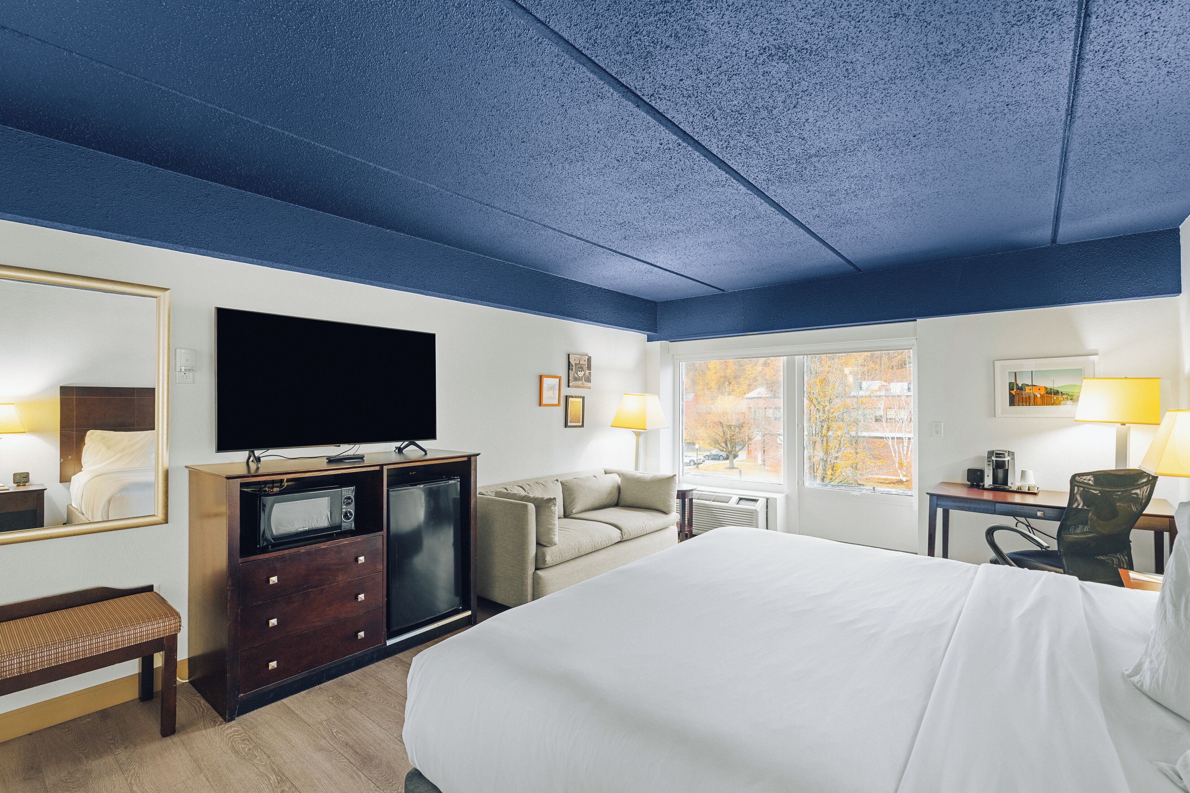 A hotel room with a large bed, a flat-screen TV, a microwave, a sofa, a desk with a lamp, and a window with a view of the outside.