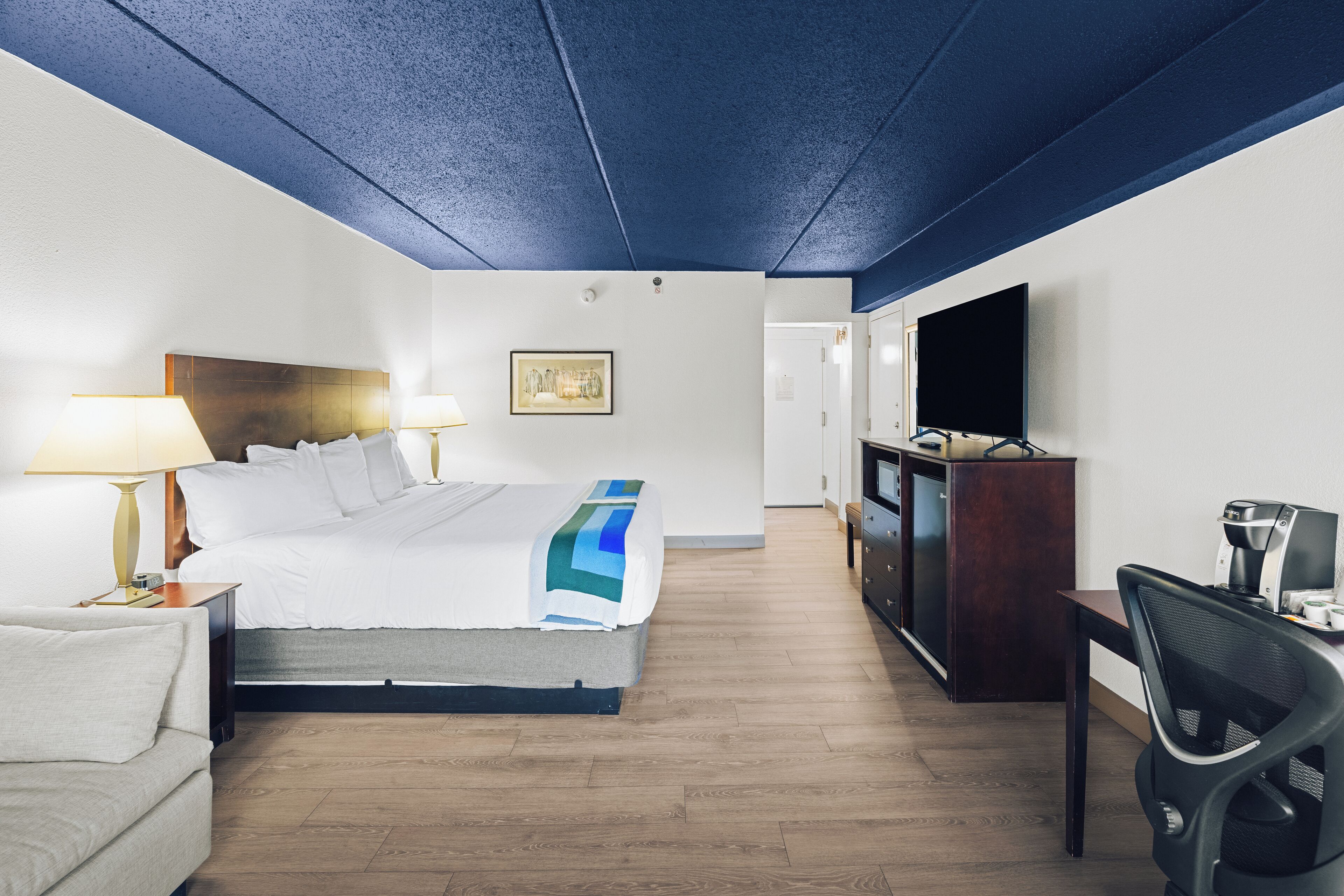 A hotel room with a bed, bedside table with lamp, desk with chair, TV, and a flat-screen TV mounted on the wall.