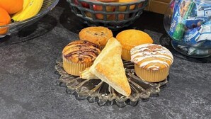 A kitchen counter with a fruit basket, a bowl of bananas, a basket of apples, muffins, and a jar of snacks.
