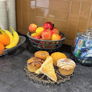 A kitchen counter with a fruit basket, a bowl of bananas, a basket of apples, muffins, and a jar of snacks.