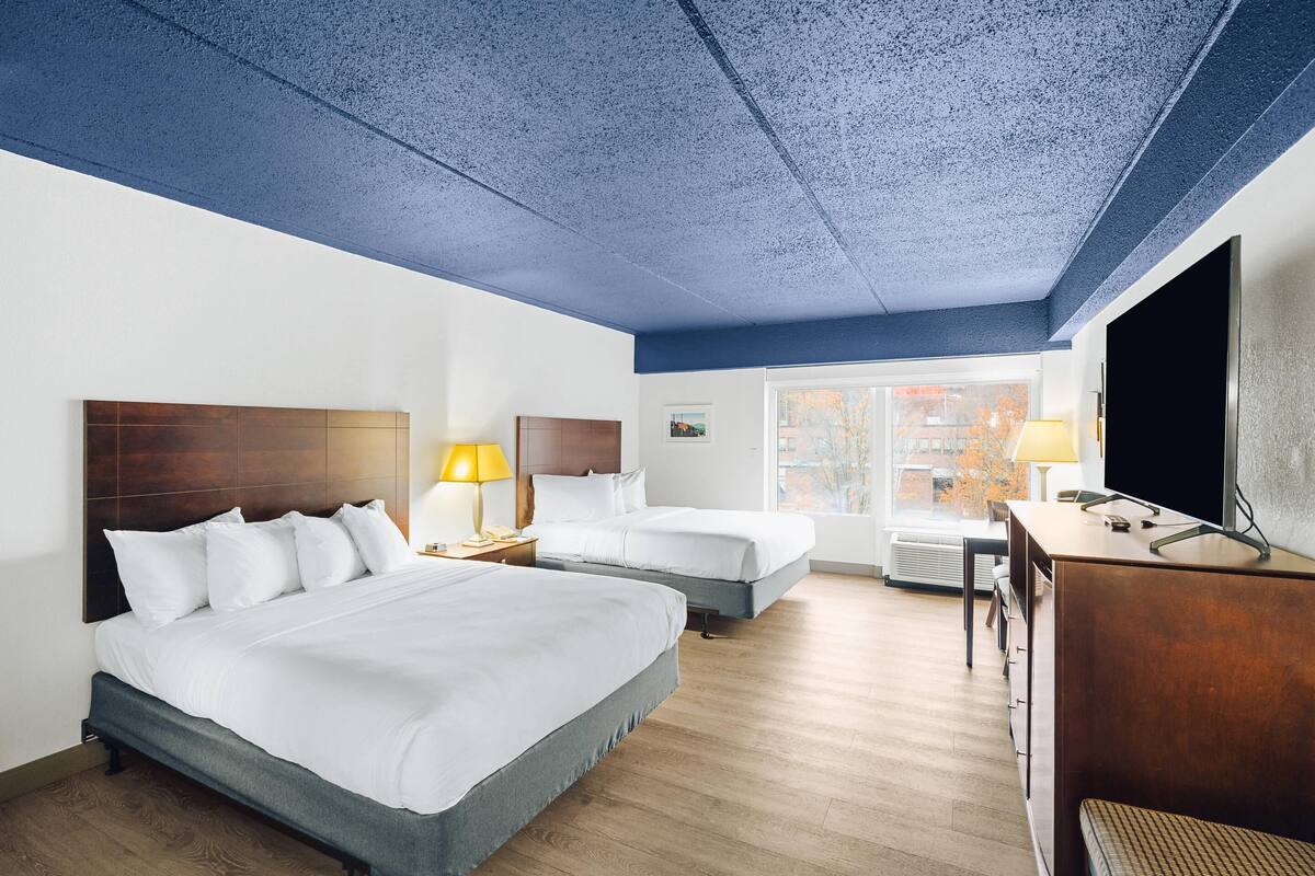 A hotel room with two beds, a desk, a TV, and a window with a view of buildings.