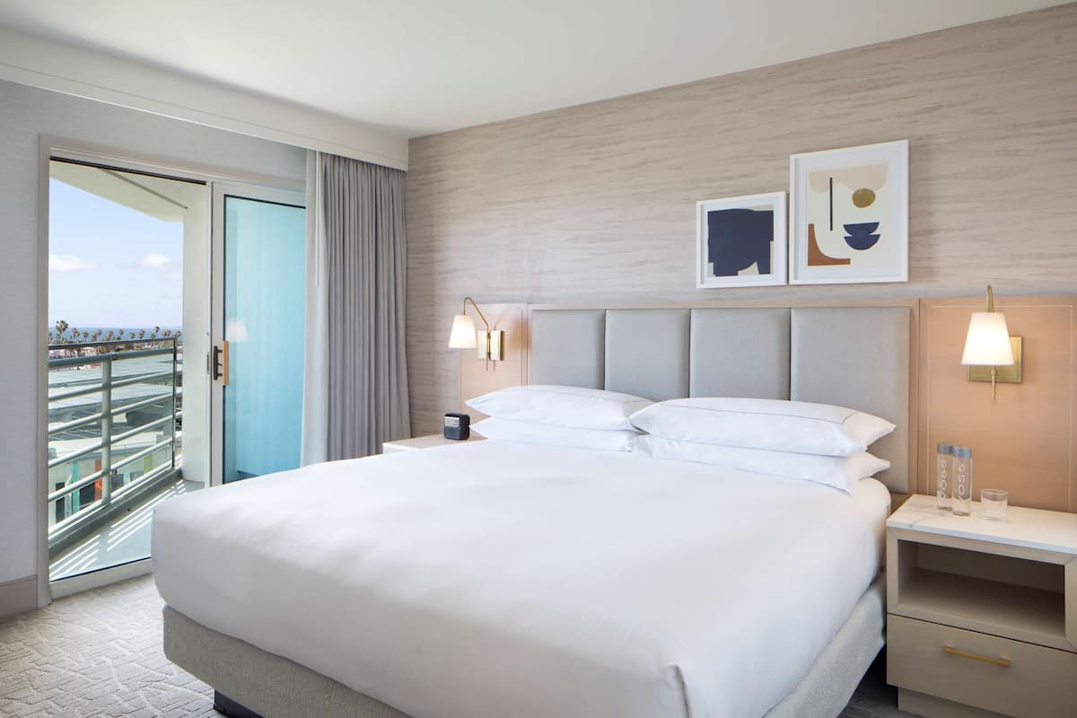 A hotel room with a large bed, a nightstand, a bedside table, a window with curtains, and artwork on the wall.