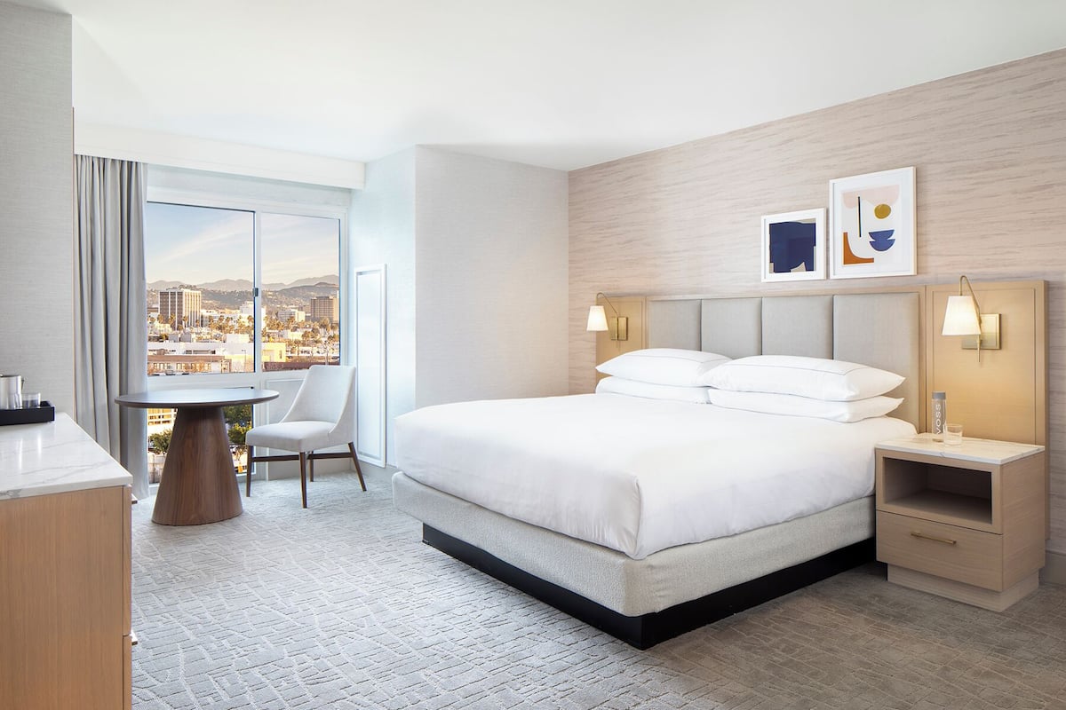 A hotel room with a bed, bedside tables, a chair, a desk, and a view of the city.