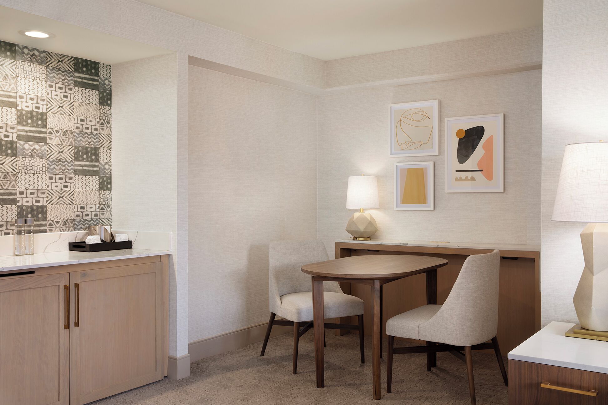 Une chambre d’hôtel avec un meuble en bois, une petite table ronde et deux chaises, une lampe fixée au mur et des œuvres d’art encadrées.
