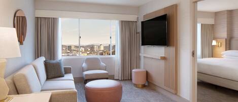 A hotel room with a sofa, armchair, ottoman, bed, TV, and a view of the city.