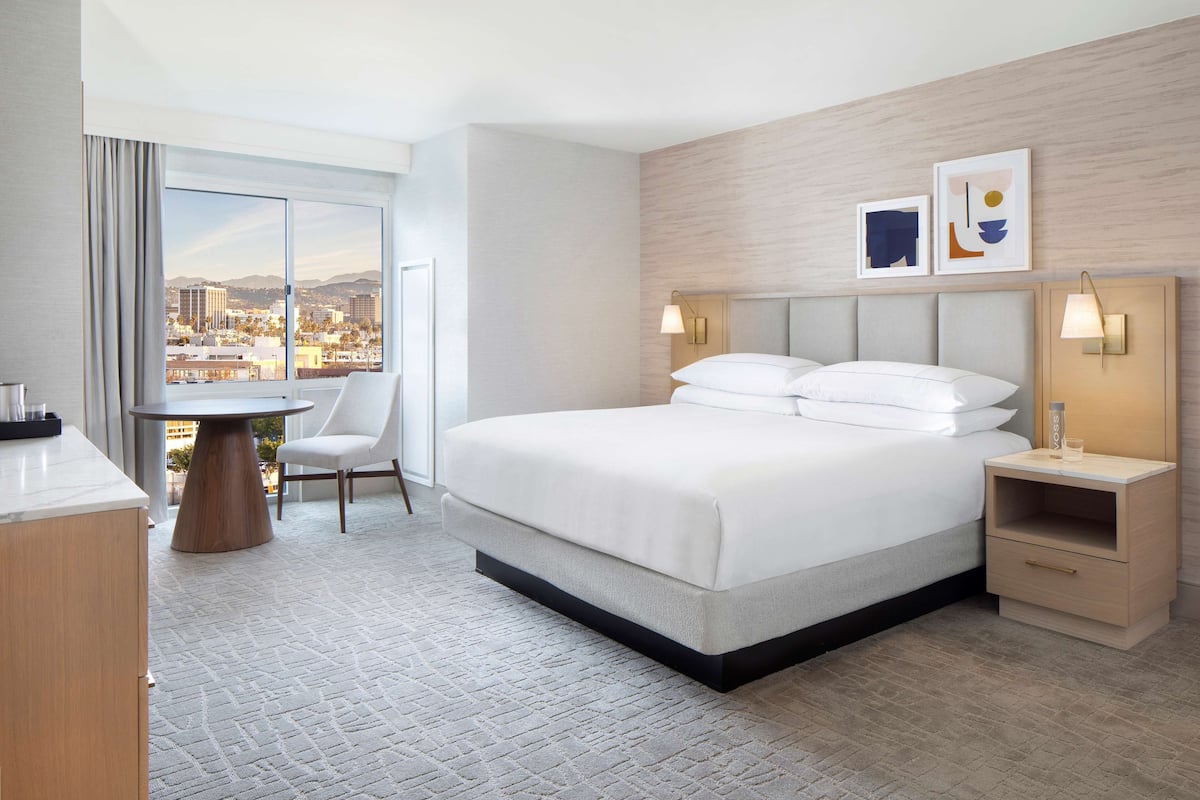 A hotel room with a large bed, a desk with a chair, a view of the city, and a nightstand with a lamp.