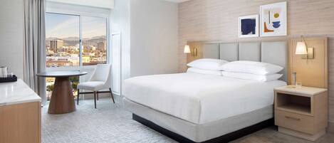 A hotel room with a large bed, a desk with a chair, a view of the city, and a nightstand with a lamp.
