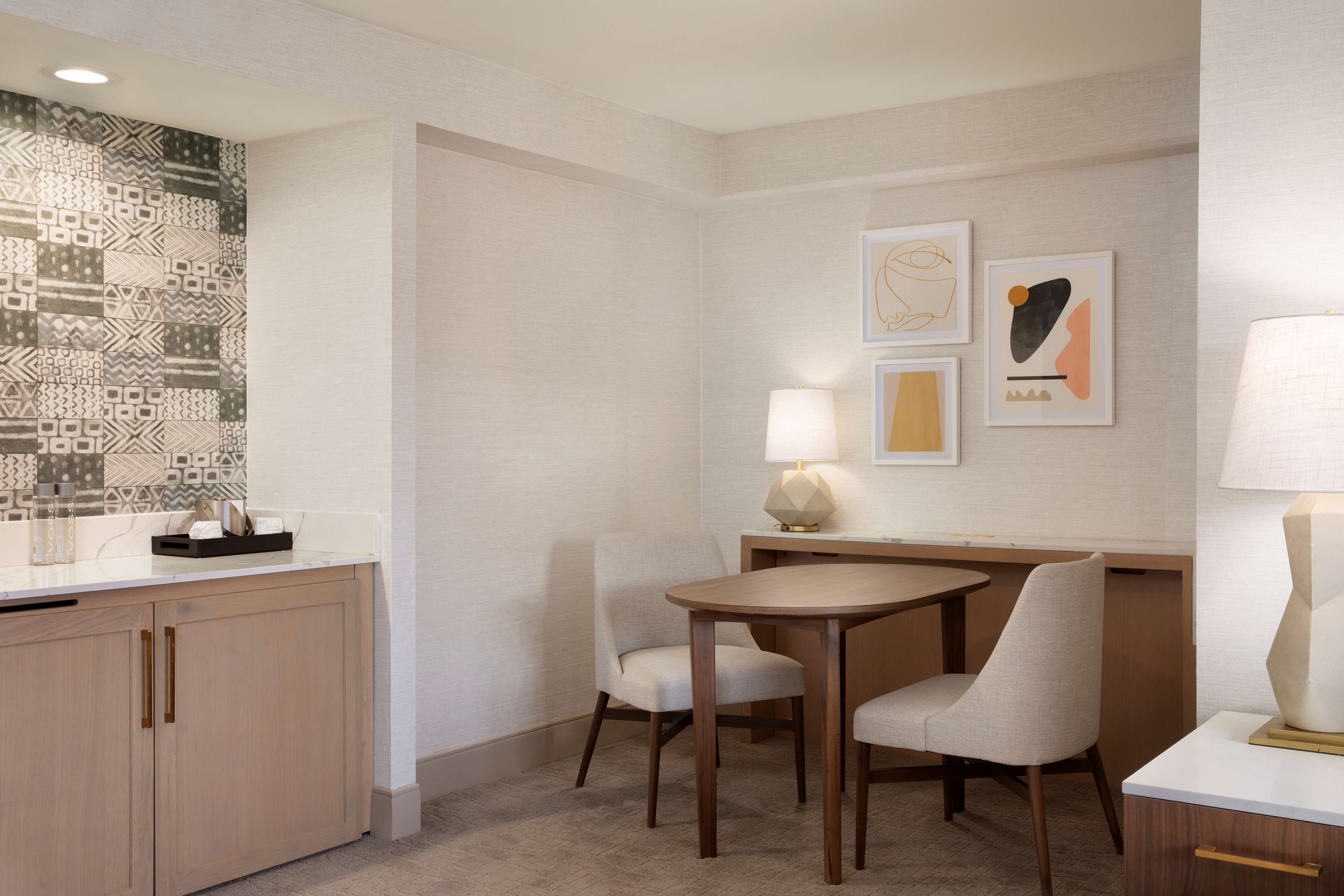 A hotel room with a dining area featuring a round table and two chairs, a small cabinet with a mirror, and a wall with patterned tiles and artwork.