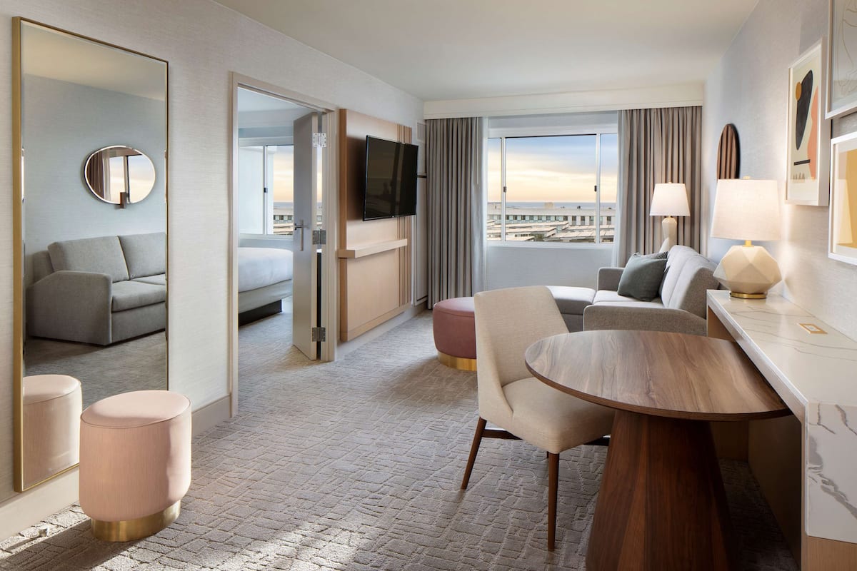 A modern hotel room with a large bed, a sitting area with a table and chairs, a television, and a view of the cityscape.