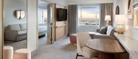 A modern hotel room with a large bed, a sitting area with a table and chairs, a television, and a view of the cityscape.