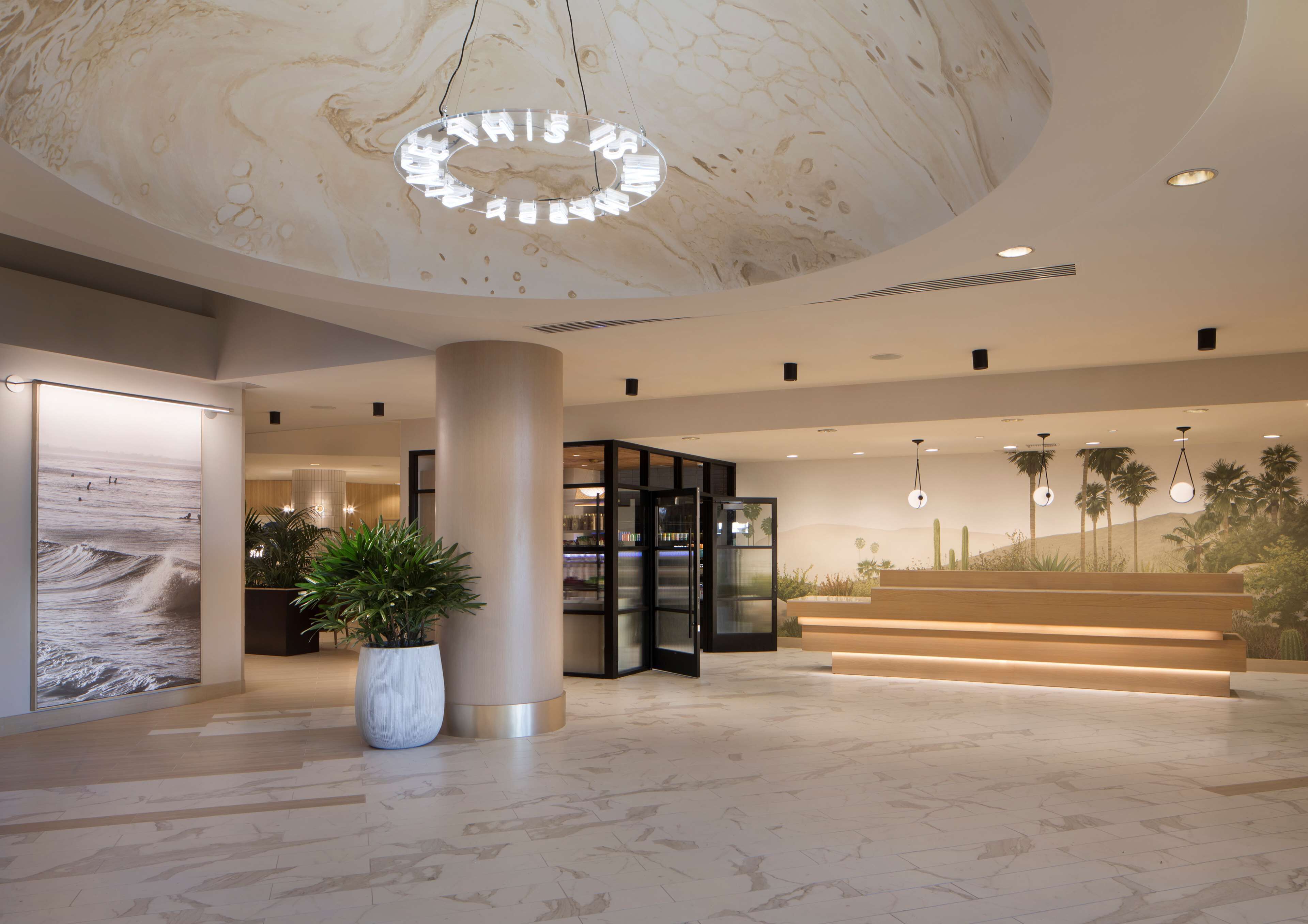 Eine moderne Lobby mit Marmor Decke, einer großen Topfpflanze, einem Glastür-Eingang und einer Treppe im Hintergrund mit Palmen.