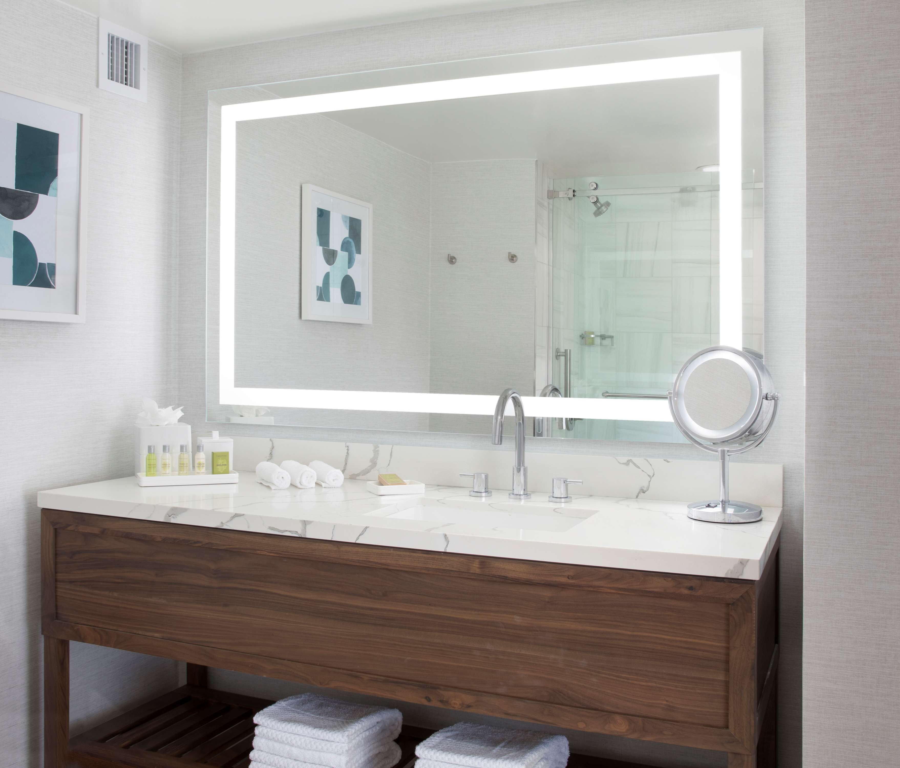 A modern bathroom with a large mirror, white marble countertop, wooden vanity, and a glass shower enclosure.