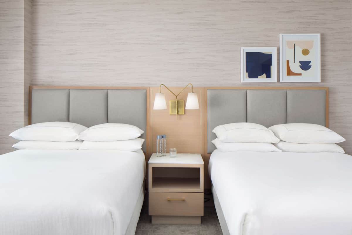A hotel room with two beds, a nightstand with a lamp, and artwork on the wall.