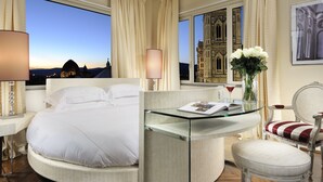 Habitación de hotel con una cama, una mesa de vidrio, una silla, un florero con flores y vista a un edificio a través de la ventana.