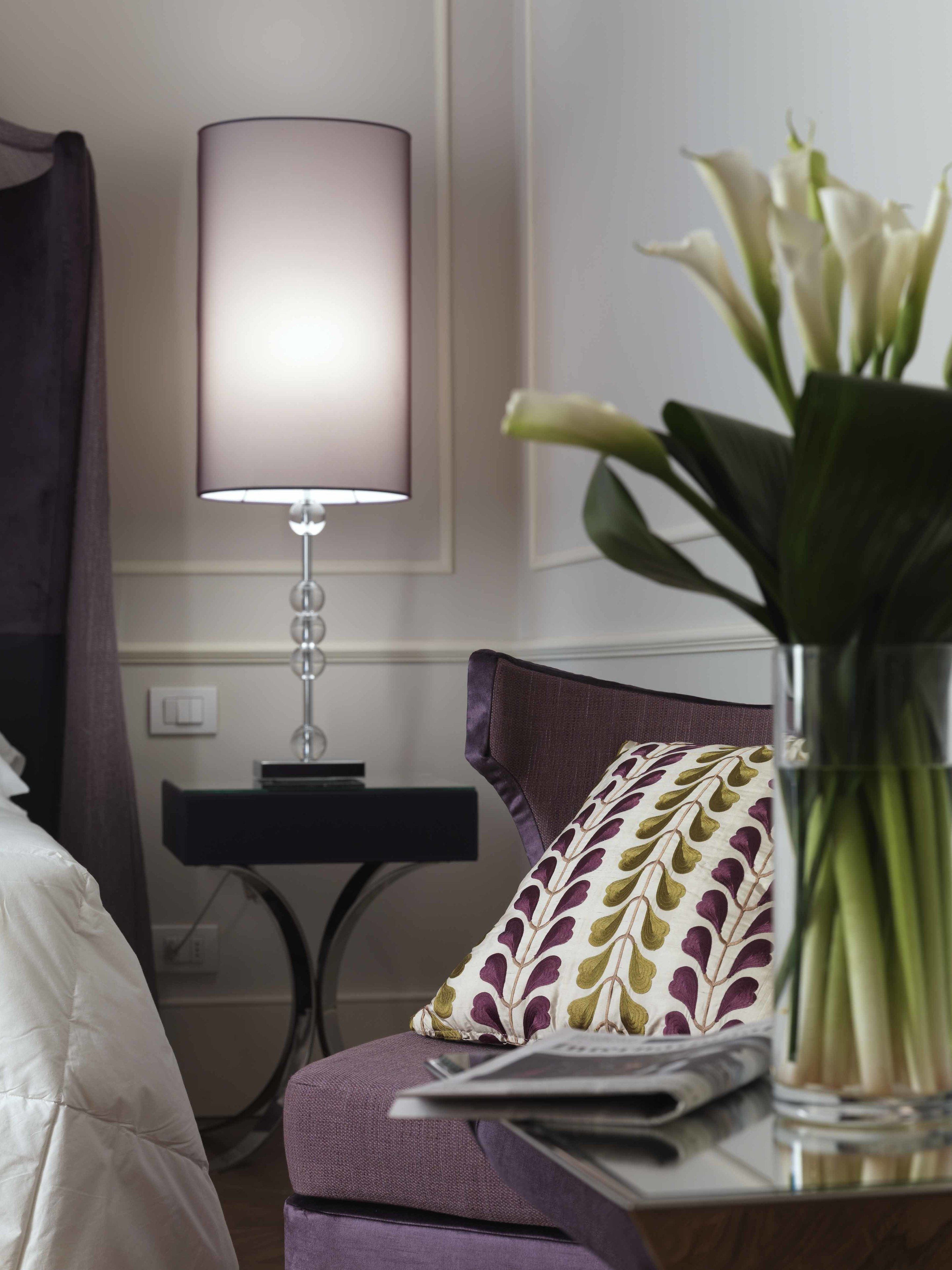 Habitación de hotel con cama, una silla con cojín estampado, una mesa con lámpara y un florero con flores.