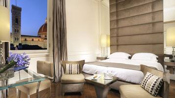 A hotel room with a large bed, two armchairs, a glass coffee table, and a view of a historic building.