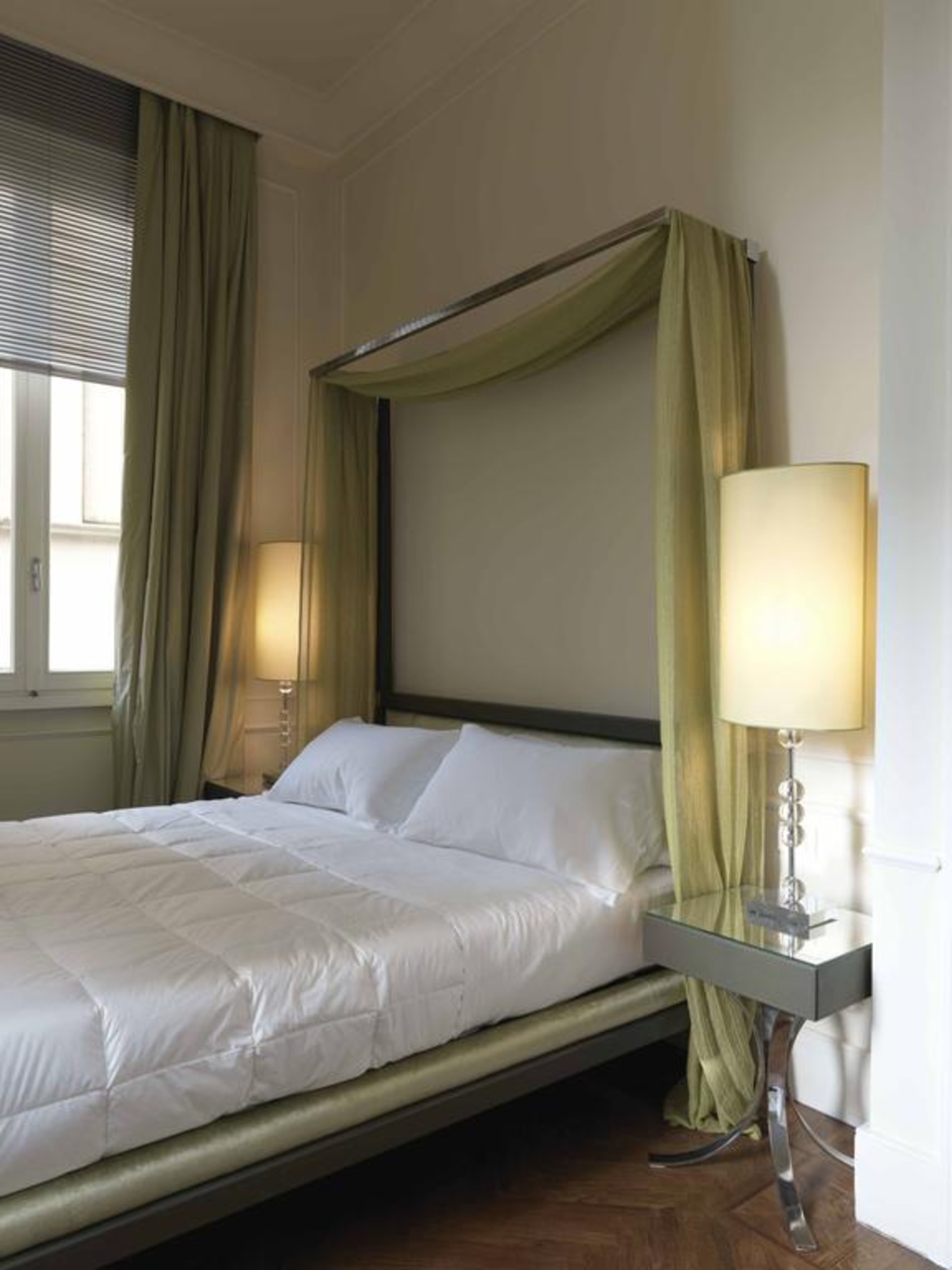 A neatly made bed with white linens, a bedside table with a lamp, and a window with curtains.