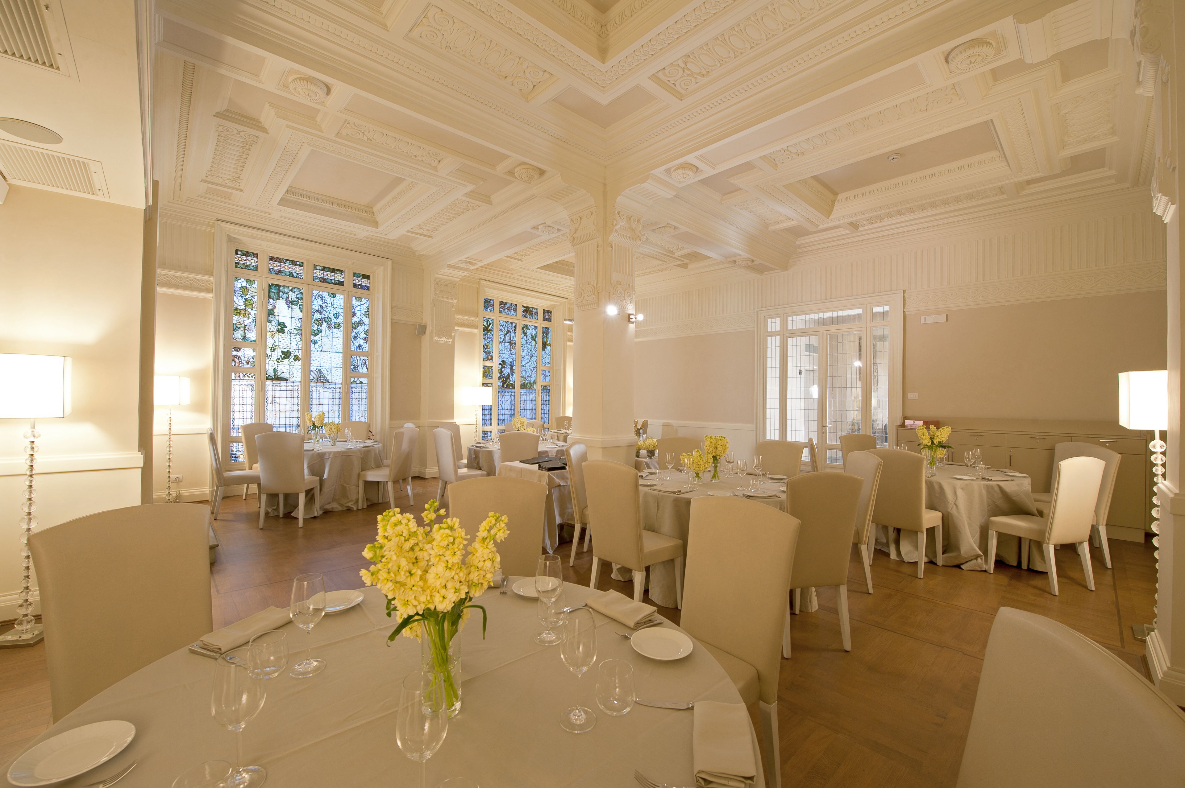A formal dining room with multiple tables set for service, large windows with decorative glass, and a classic ceiling design.
