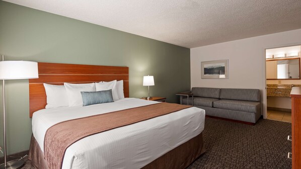 A hotel room with a large bed, a bedside table with a lamp, a grey sofa, a wooden headboard, and a framed picture on the wall. - Best Western Buffalo Ridge Inn (Custer)