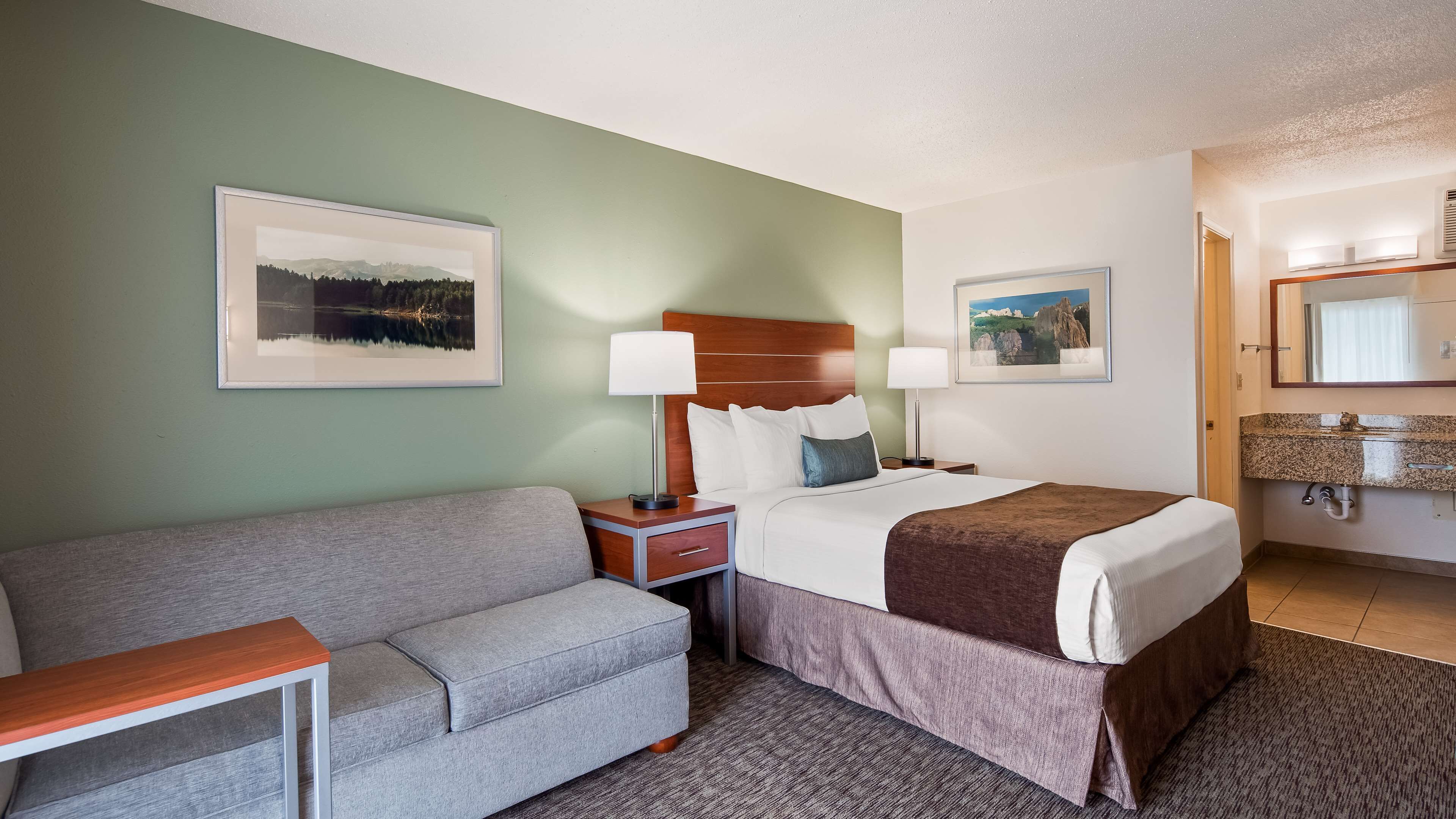 Habitación de hotel con cama, mesita de noche con lámpara, sofá, pequeña mesa y baño con lavabo y espejo.