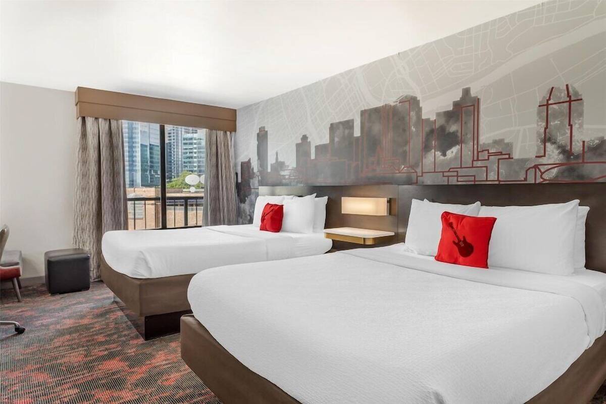 A hotel room with two beds, a cityscape mural, a view of buildings, and a large window with curtains.