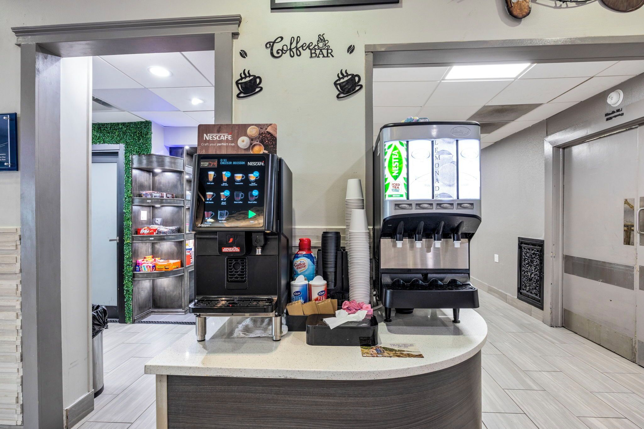 A coffee bar with a Nescafe machine and a KELLS machine, surrounded by stacks of disposable cups and a trash bin.