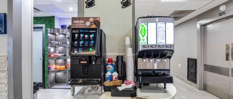 A coffee bar with a Nescafe machine and a KELLS machine, surrounded by stacks of disposable cups and a trash bin.