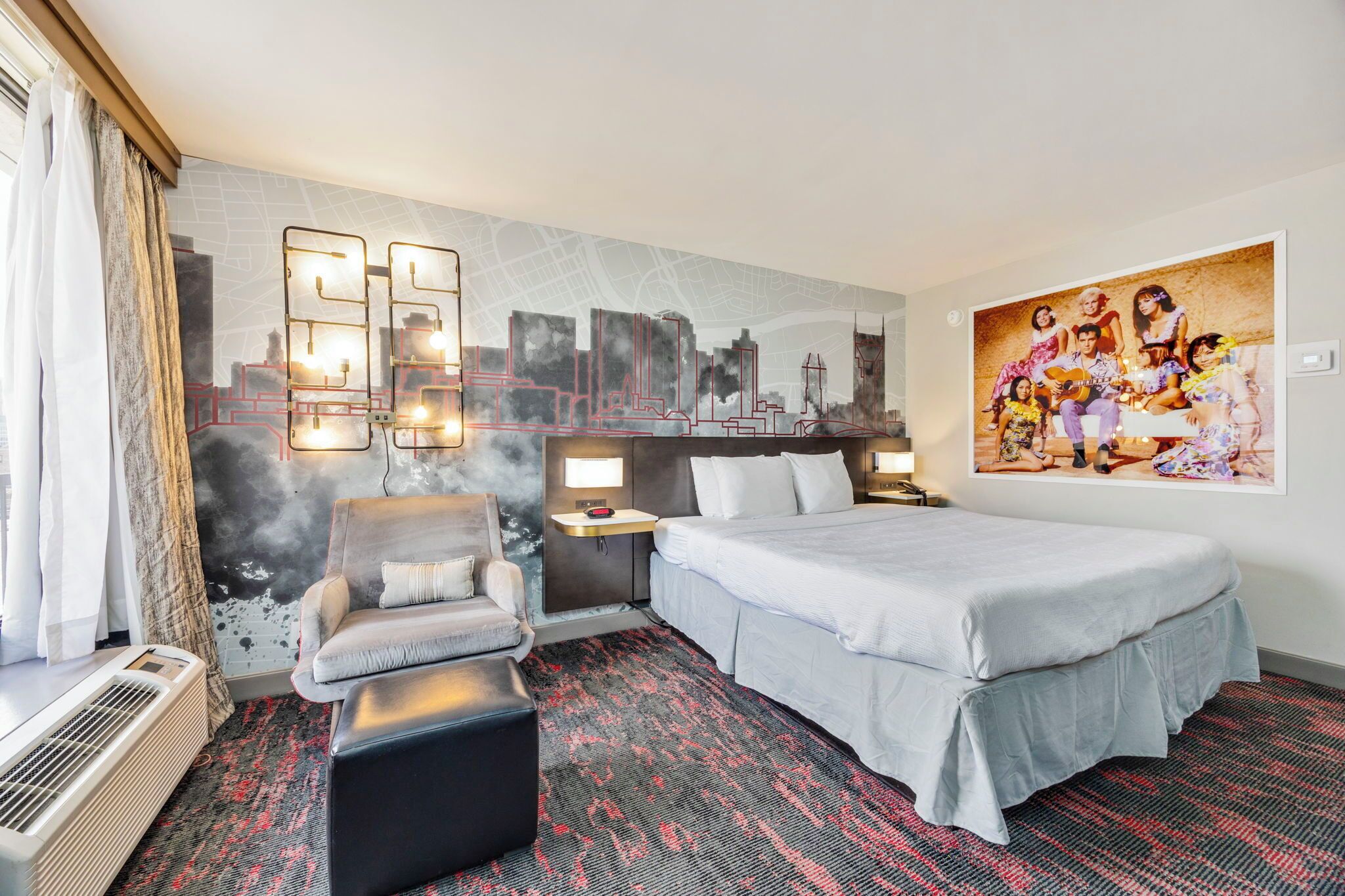 A hotel room with a bed, a chair, a small table, and a wall with a cityscape and a family portrait.