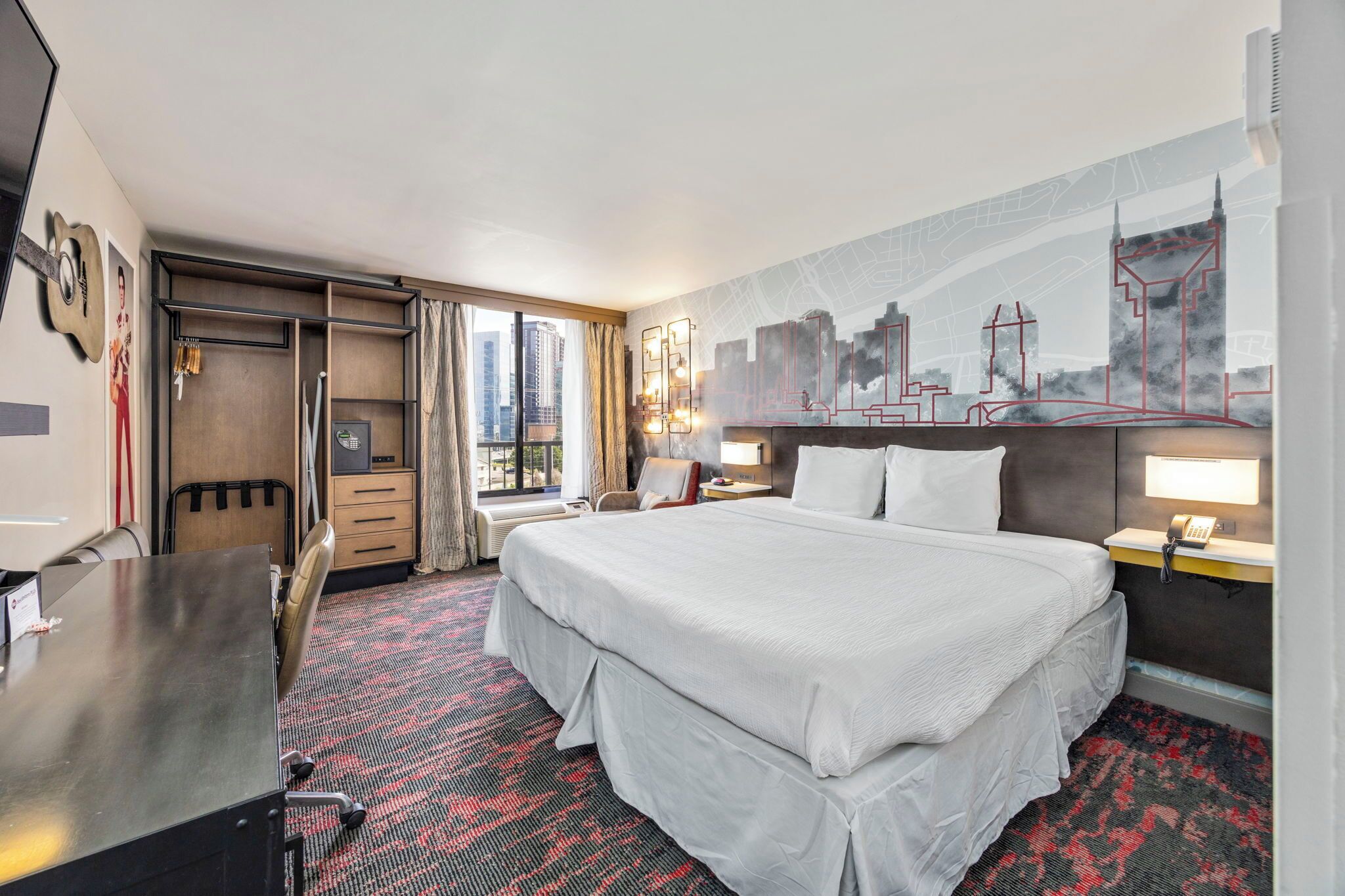 A hotel room with a large bed, a desk, a chair, a wardrobe, and a cityscape mural on the wall.