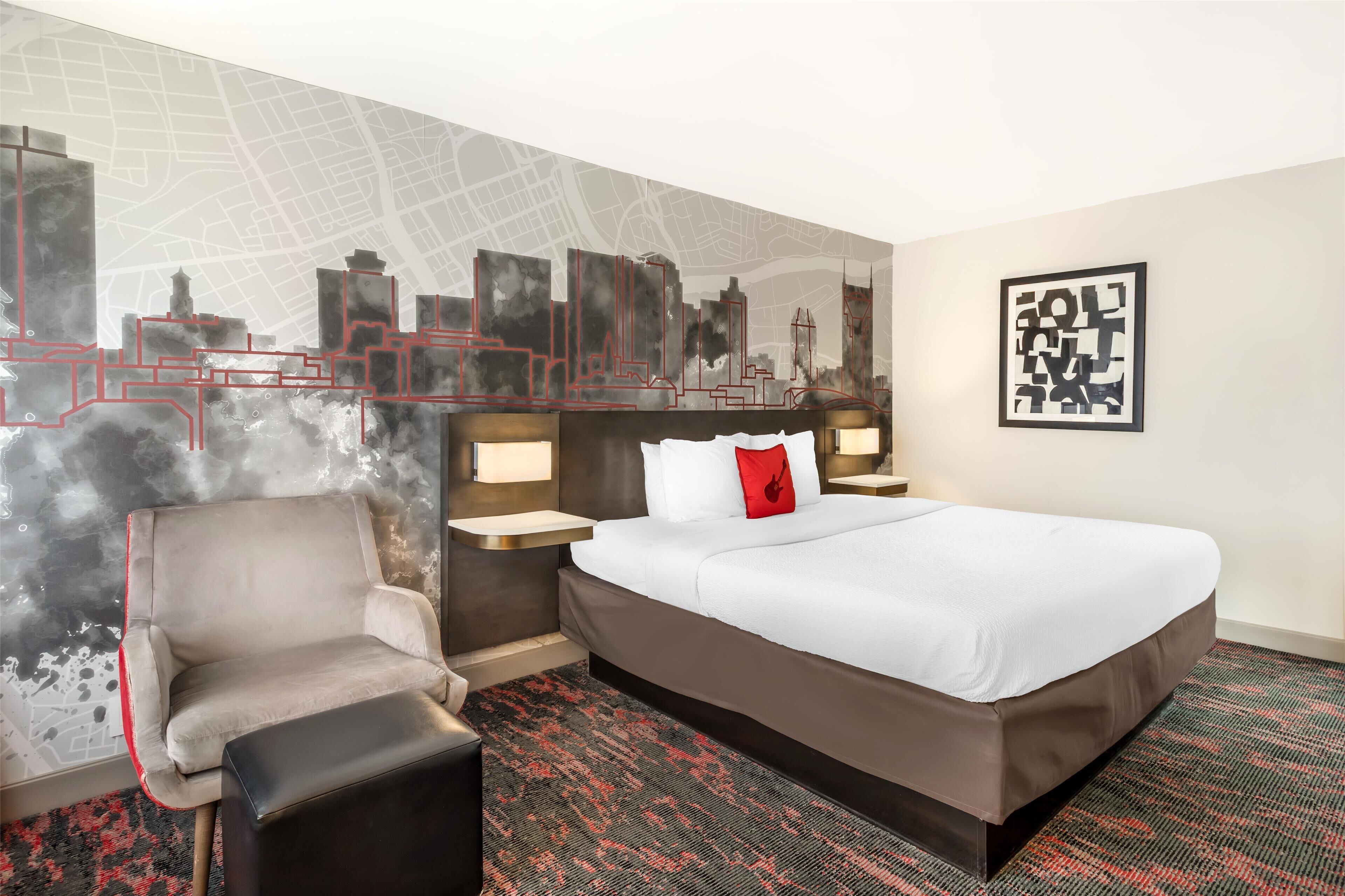 A hotel room with a large bed, a chair, a nightstand, and a wall with a cityscape design.