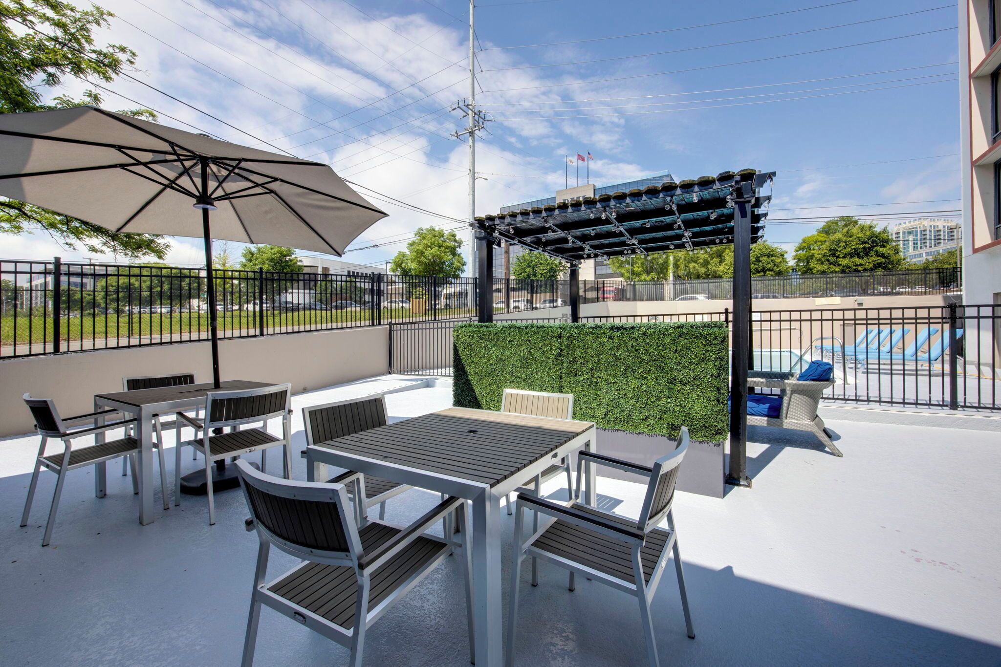 An outdoor patio with tables and chairs, a pergola, and a view of a swimming pool and buildings in the background.