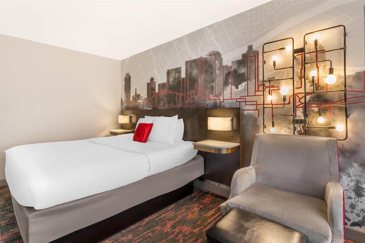 A hotel room with a large bed, a grey armchair, a cityscape mural, and modern lighting fixtures.