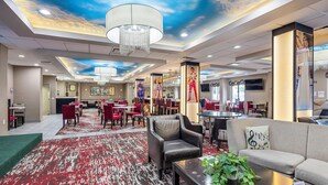 A hotel lobby with a red and white patterned carpet, a chandelier, a sofa with a musical note pillow, and a bar area with red chairs.