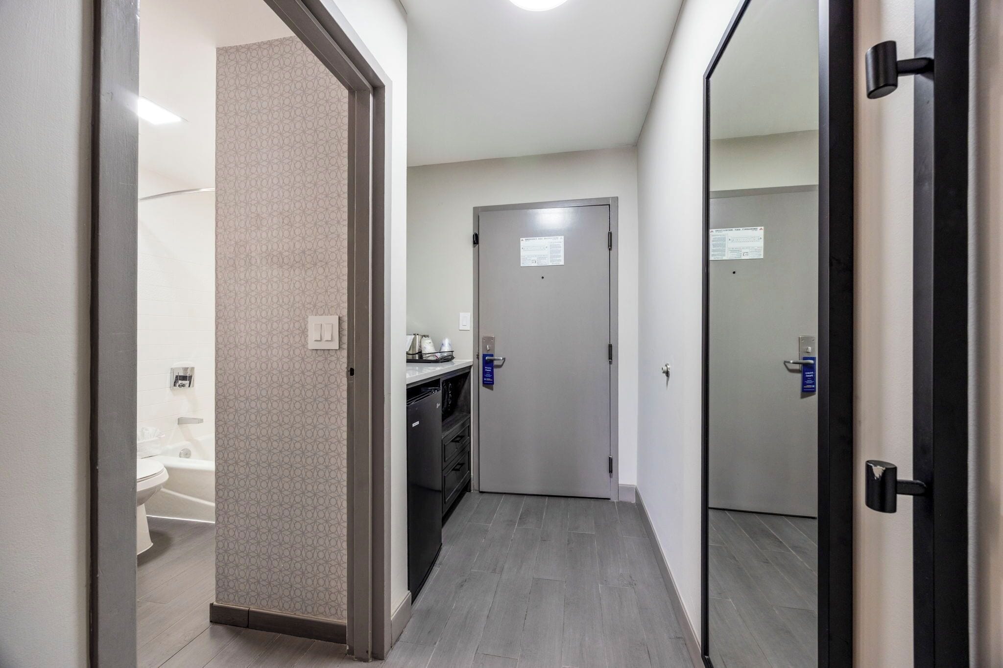 Un couloir étroit avec une porte donnant accès à une salle de bain, à un coin cuisine et à un mur recouvert de miroirs.