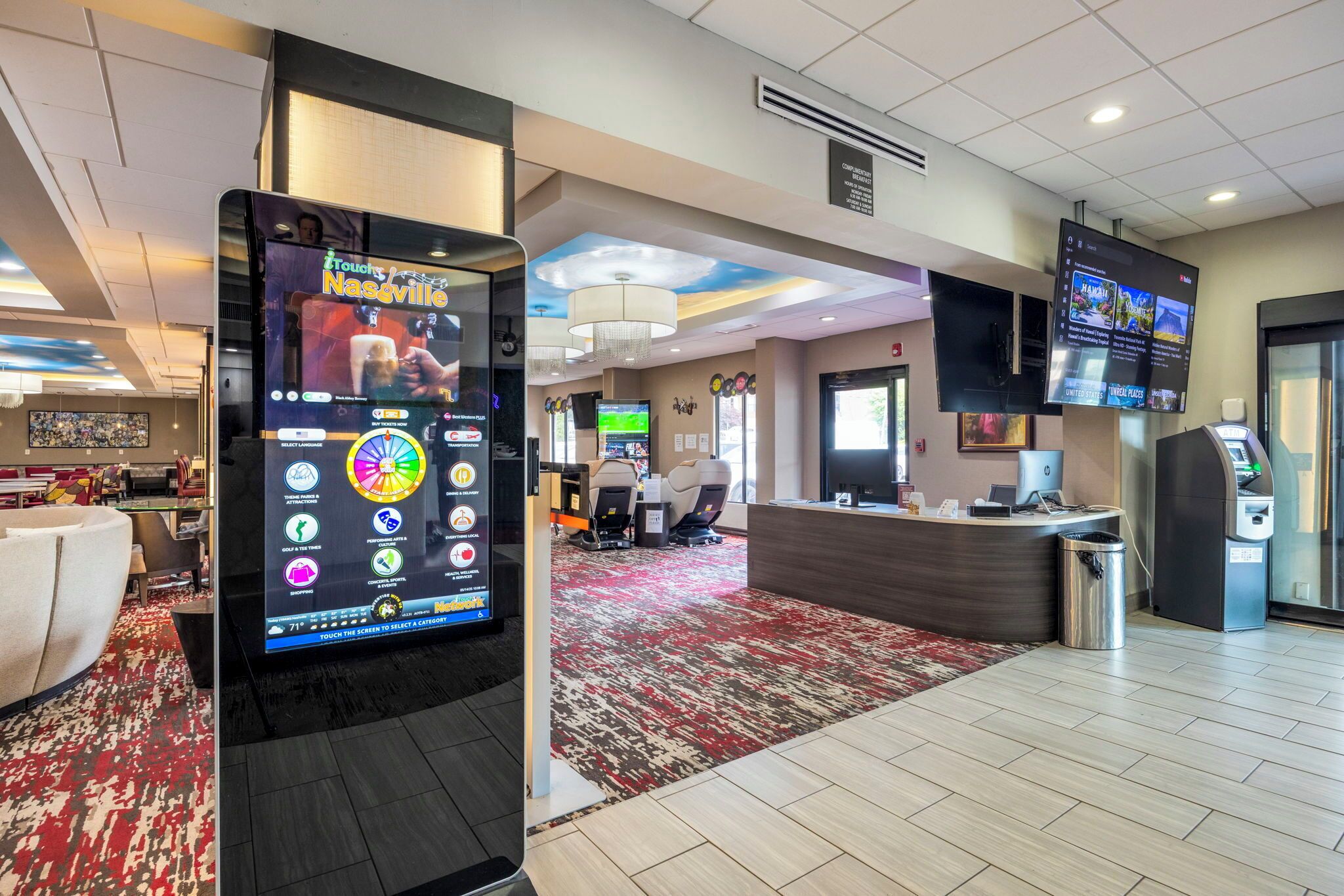 Vestíbulo de hotel con quiosco digital, zona de estar, mostrador de recepción y una pantalla grande que muestra contenido.