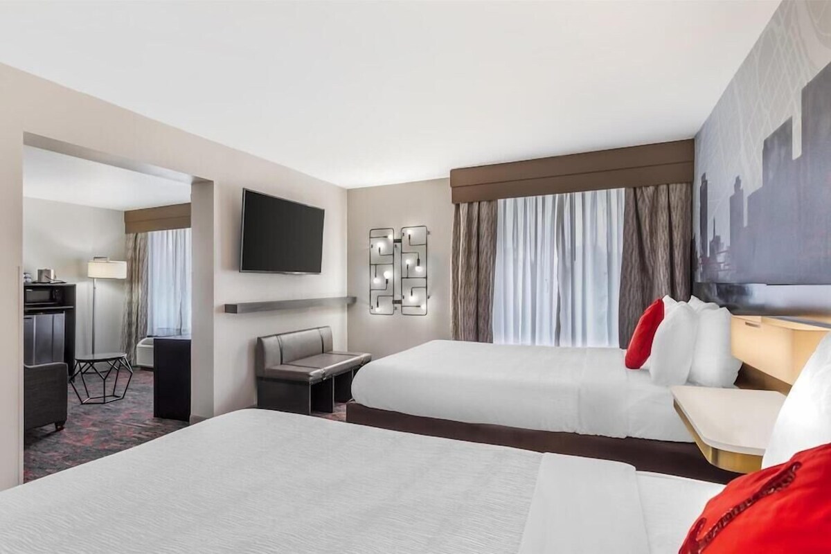 Quarto de hotel com duas camas, uma escrivaninha, uma cadeira, televisão e uma janela grande com cortinas.