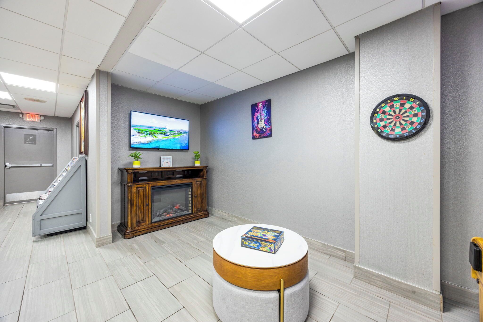 A modern lobby with a flat-screen TV, a dartboard, a painting, and a small round table with a book.