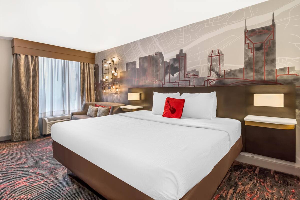 A hotel room with a large bed, a cityscape mural, a desk with a lamp, and a window with curtains.