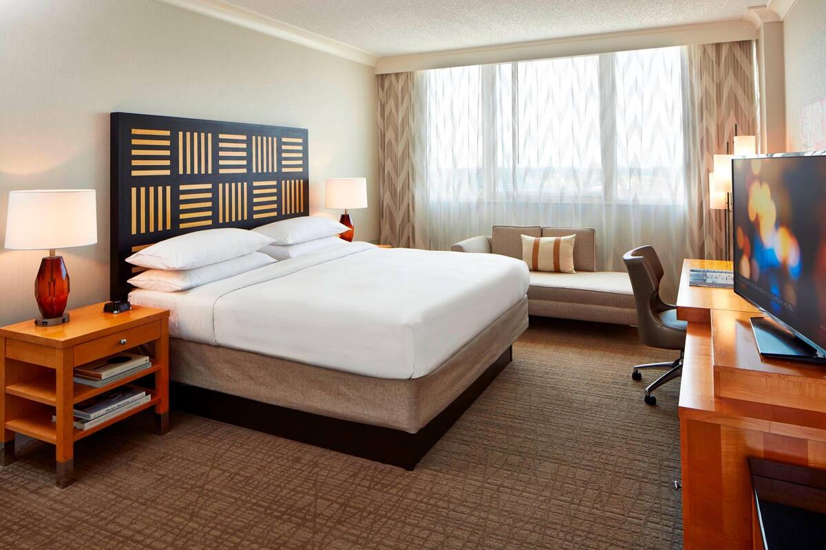 A hotel room with a large bed, a desk with a flat-screen TV, a sofa, and a window with sheer curtains.