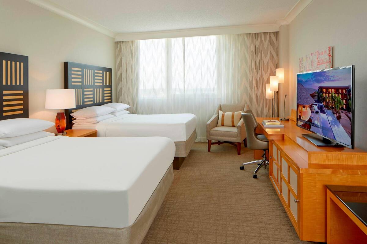 A hotel room with two beds, a desk with a TV, a chair, and a large window with curtains.