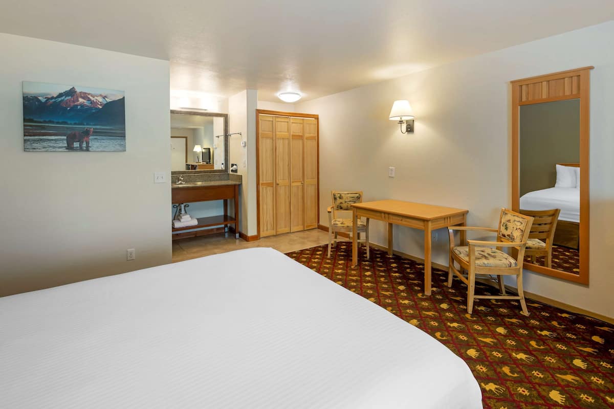 A hotel room with a large bed, a wooden desk, two chairs, a mirror, and a painting of a mountain scene.