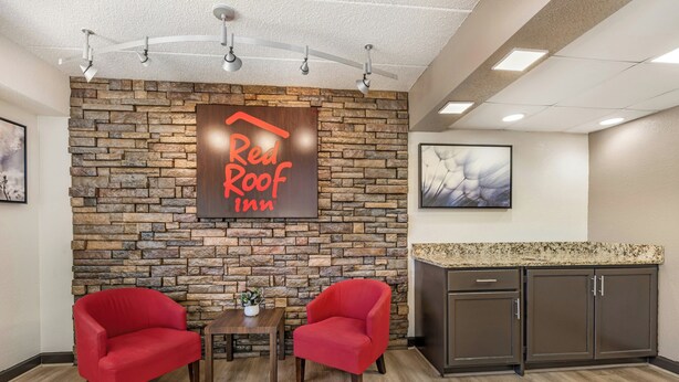 A Red Roof Inn lobby with a stone wall, two red armchairs, a small table with a plant, and a kitchen area with cabinets. - Red Roof Inn Chicago - Joliet (Joliet)