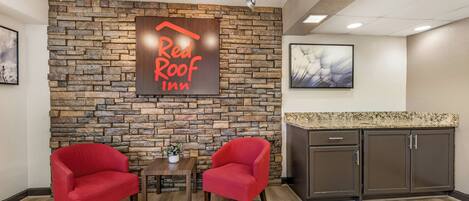 Vestíbulo de Red Roof Inn con pared de piedra, dos sillones rojos, una mesa pequeña con una planta y una zona de cocina con armarios.